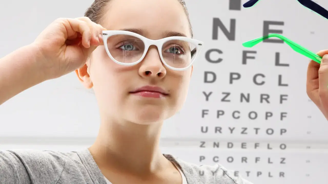 Dziewczynka z wadą wzroku - 20, przymierza okulary na tle tablicy Snellena.
