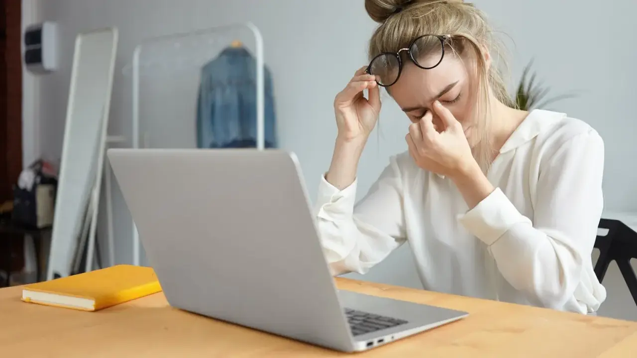 Zmęczona kobieta z okularami masuje oczy, siedząc przy laptopie. Czas zadbać o to, jak chronić oczy przed komputerem.