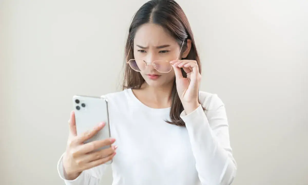 Kobieta z okularami próbuje odczytać coś na telefonie, marszcząc brwi. Może to być objaw astygmatyzmu krótkowzrocznego.