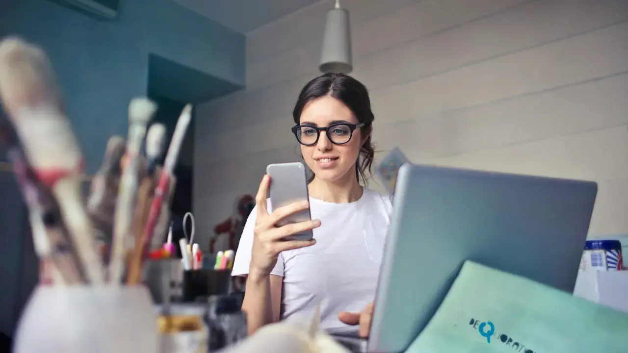 Kobieta w okularach do komputera przegląda telefon, siedząc przy laptopie.