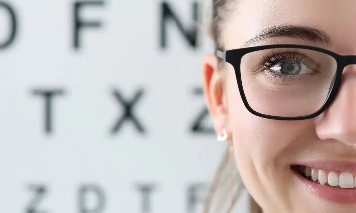 Uśmiechnięta kobieta w okularach na tle tablicy Snellena. Wizyta u optometrysty lub okulisty to ważny krok do dobrego widzenia.