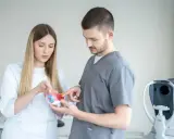 Lekarze omawiają model oka, zastanawiając się, czy wadę wzroku da się wyleczyć. W tle aparat do badania wzroku.