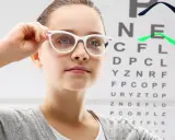 Dziewczynka z wadą wzroku - 20, przymierza okulary na tle tablicy Snellena.