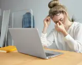 Zmęczona kobieta z okularami masuje oczy, siedząc przy laptopie. Czas zadbać o to, jak chronić oczy przed komputerem.