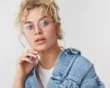 Młoda kobieta z kręconymi blond włosami i okrągłymi okularami, jej twarz w kształcie serca jest delikatnie podparta dłonią.
