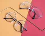 Dwa stylowe okulary, okrągłe i aviator, leżą na pomarańczowo-czerwonym tle. Złoty łańcuszek oplata je, sugerując, że to okulary korekcyjne, być może na astygmatyzm.