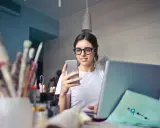 Kobieta w okularach do komputera przegląda telefon, siedząc przy laptopie.