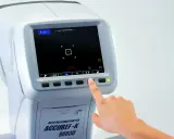 Dłoń naciska przycisk na auto refraktometrze, pokazując ekran z danymi badania wzroku. Pozwala to zrozumieć, jak odczytać wydruk z komputerowego badania wzroku.