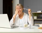 Zmęczona kobieta pociera oko, patrząc na ekran laptopa, który emituje niebieskie światło. Obok leżą okulary i słuchawki.