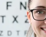 Uśmiechnięta kobieta w okularach na tle tablicy Snellena. Wizyta u optometrysty lub okulisty to ważny krok do dobrego widzenia.
